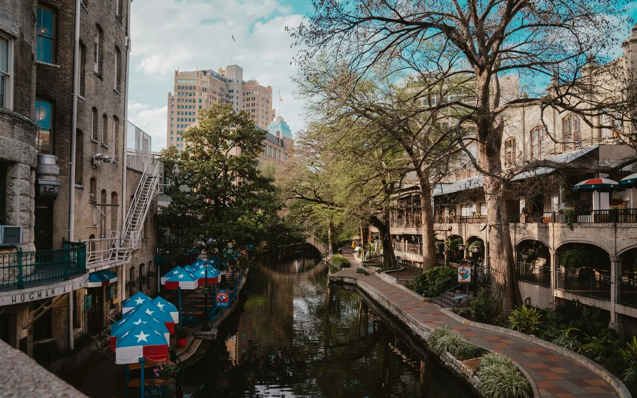 The San Antonio River Walk by day: the river between stone walkways, cypress trees, and cafe umbrellas, with downtown buildings beyond